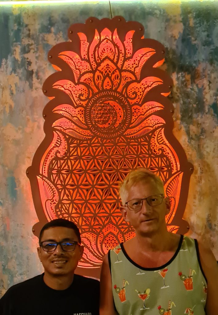 Zwei Männer, ein dunkelhaariger Thai und ein blonder Europäer, lächeln vor einer roten Ornamentwand in die Kamera